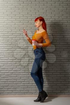 Young red-haired girl with a cup of coffee is reading something thoughtfully on her smartphone against the background of a gray brick wall