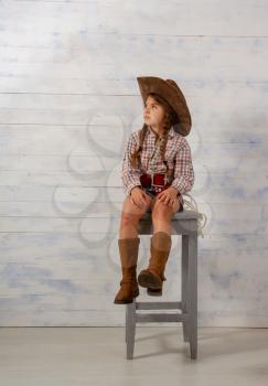 Little girl in a wide-brimmed cowboy hat and traditional dress in high boots and with a lasso posing on a light wooden background