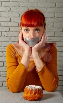Conceptual image of a young red-haired girl on a diet sitting with her mouth taped with silver tape before appetizing pastries