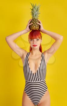 A young girl in a retro striped bathing suit put a pineapple on her head on a bright yellow background