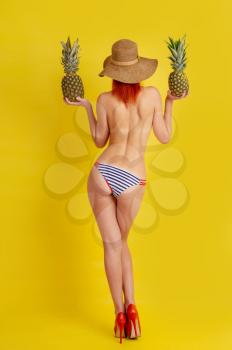 Young semi-naked girl in retro striped bathing shorts and high-heeled shoes and a wide-brimmed hat holds two pineapples in her hands while standing with her back to the viewer on yellow background.