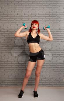 Young slim girl in sportswear is engaged in training and lifts dumbbells against a gray brick wall