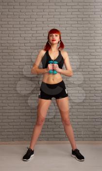 Young slim girl in sportswear is engaged in training and lifts dumbbells against a gray brick wall