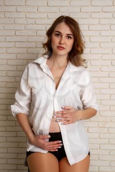 A beautiful young girl in her sixth month of pregnancy in a shirt of her husband and black dignified golf sits on a rough high stool against a brick wall.