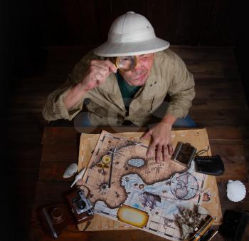 An adult explorer in a cork helmet over an old wooden table paves a route using old maps