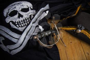 Classic pirate black felt captain's cocked hat and old sword lying on a wooden floor next to the Jolly Roger flag
