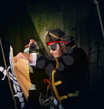 Adult pirate captain in a traditional costume and with arms looks at a treasure map against the backdrop of a jolly roger