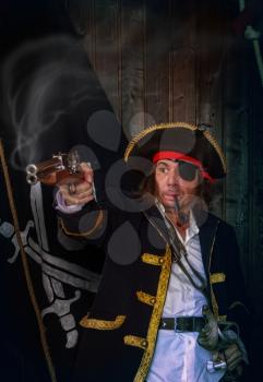 Adult pirate captain in a traditional costume shoots a pistol and holds a sword in his hands against the background of a jolly roger