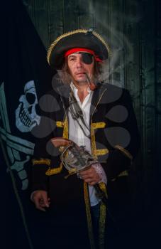 Adult pirate captain in a traditional costume and with a weapon smokes a pipe against the background of a jolly roger
