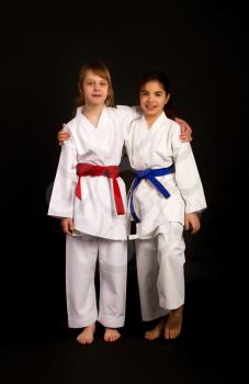 Two little karate girl friends in white kimonos, one in red and the other in blue competition belts hugging against a dark background