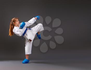 Little girl in a white kimono with a blue belt and equipment carries out blows on a dark background