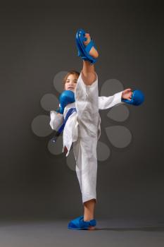 Little girl in a white kimono with a blue belt and equipment carries out blows on a dark background