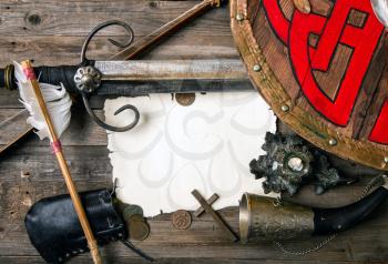 Vintage medieval background with sword and shield coins and other knightly things blank sheet of paper blank for writing or card on a rough wooden table