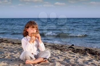 beautiful little girl sitting on beach and play pan pipe