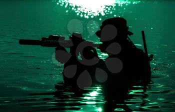 Silhouette of special forces with rifle in action during night raid crossing river in the jungle waist deep in the water. Moon path glowing
