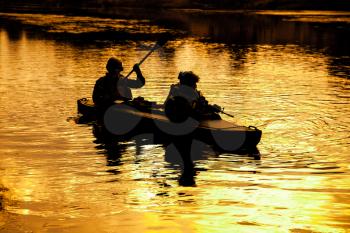 Silhouette of special forces men paddling army kayak. Boat moving calmly across the river, diversionary mission, sunset dusk