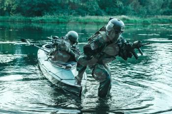 Covert landing. Pair of armed operators with painted faces disembarking river coast from military kayak . Diversionary mission under cover of darkness
