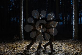 Black silhouettes of pair of soldiers in the forest moving in battle operation. Back light
