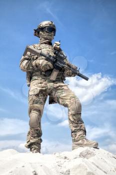 Man in military camouflage uniform and mask, equipped tactical ammunition, standing on sand dune with service rifle replica in hands, cloudy sky on background. Airsoft player taking part in war games