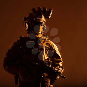 Army elite forces member, modern infantryman with hidden face, in tactical ammunition, equipped radio headset, night vision device mounted on helmet, standing with short barrel service rifle in hands