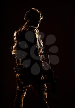 Low key studio shoot of army special forces soldier, commando fighter in mask, ballistic glasses, tactical helmet and battle uniform, holding short barrel service rifle, looking back over shoulder