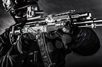 Close up photo of modern assault rifle with collimator sight in hands of police special operations group fighter in black uniform, helmet and face hidden behind mask, desaturated, low key studio shoot