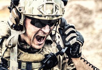 Special forces soldier, military communications operator or maintainer in helmet and glasses, screaming in radio during battle in desert. Calling up reinforcements, reporting situation on battlefield