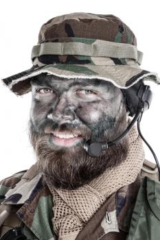 Shoulder studio portrait of commando soldier, modern mercenary, professional soldier with black camouflage paint on bearded face, tactical radio headset with microphone, isolated on white background