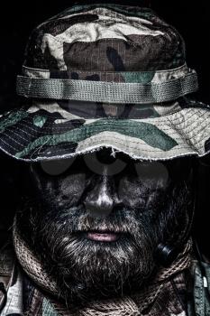 Close up, low key portrait of bearded commando fighter, army special forces soldier, private military company mercenary in boonie hat, tactical radio headset, black and green face camouflage paint