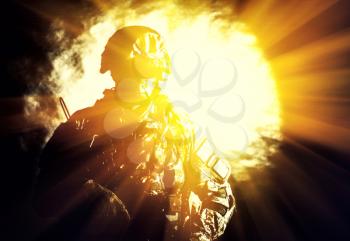 Low key studio portrait of private security service contractor, army infantry rifleman, US marine raider in blinding light of projector or searchlight in helmet, sunglasses, camouflage uniform posing with weapon on black background with backlight