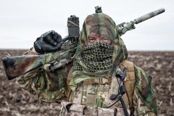 Army marksman, airsoft player in camouflage uniform and load carrier, masking cape on head, armed service rifle with optical sight, hiding face with shemagh, standing on field, looking into distance