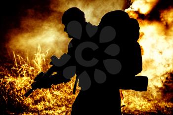 Silhouette of army soldier, elite troops fighter armed service rifle, carrying tactical backpack on back, standing on background of fiery exposure. Combatant moving in darkness on burning battlefield