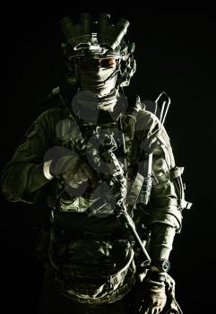 Army elite troops serviceman, counter-terrorist team member wearing mask and glasses, equipped night-vision device, radio headset mounted on combat helmet, armed submachine gun, standing in darkness