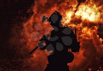 Silhouette of modern infantry soldier, elite army fighter in tactical ammunition and helmet, standing with assault service rifle in hands on background of fiery explode. Burning fire of war conflict