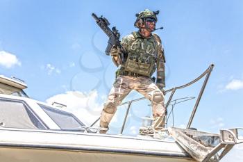 Army special forces soldier, commando fighter in full ammunition, wearing body armor and helmet, armed service rifle, standing on bow of speed boat, looking into distance during seacoast patrolling
