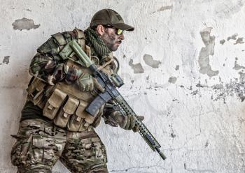 Army soldier, special operations forces rifleman in camouflage uniform, load carrier, glasses and cap standing near chipped wall with service rifle in hands. Commando fighter, combatant at war zone