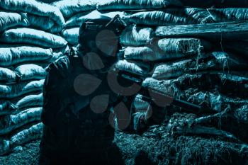Navy SEALs fighter, army soldier in combat uniform, goggles and battle helmet at night watch, sitting with assault rifle in trench with sandbags, looking in loophole, waiting for enemy attack