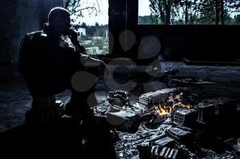 Special forces soldier after the fight sitting by the fire in ruined building smoking cigarette