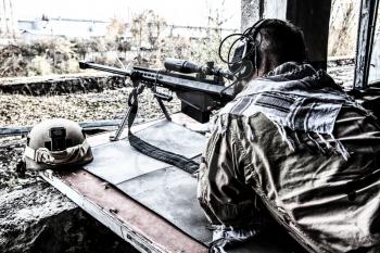 Portrait of U.S. Navy SEAL sniper on firing position, armed with large caliber sniper rifle with telescopic sight, wearing tactical headset with microphone, observing territory, searching targets