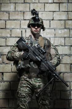 US Army Ranger with machinegun and night vision goggles standing near the wall. Front view portrait