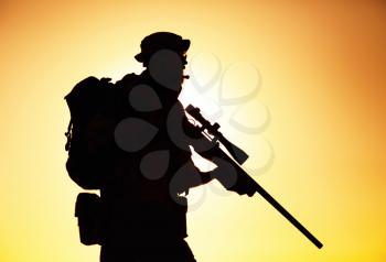 Silhouette of army elite forces sniper, commando shooter in boonie hat, carrying backpack, equipped tactical radio, walking on background of setting or raising sun. Counter terrorist forces soldier