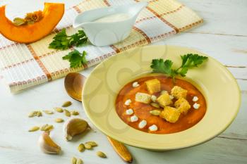 Roasted pumpkin squash vegetable soup with cream, pumpkin seeds, garlic croutons and parsley in a light yellow plate on white wooden background