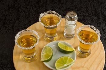 Three shots of gold tequila with lime and salt on the black background. Tequila shot. Gold Mexican tequila. Tequila