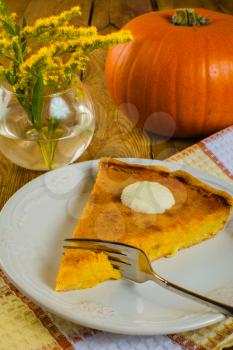 Pumpkin pie with whipped cream, a fork, a plate of white, checkered napkin, glass vase with yellow mimosa flowers on a wooden background
