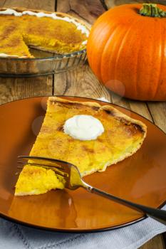 pumpkin pie slice with whipped cream on a brown plate, fork, linen napkin, glass baking dish on a wooden background