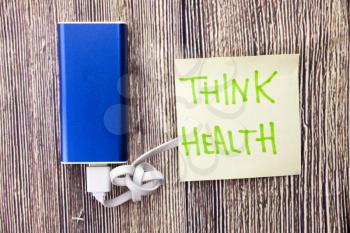 Power bank and note with the words Think Health are present on a woody surface. state of physical, mental and social well-being is mentioned in the image