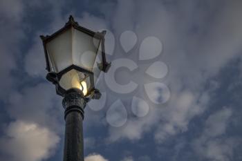 An incandescent bulb with a broken glass glowing in antique street lamp post.The medieval european lamp post is made of cast steel is made of an ornate top.