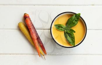 Top view of carrot soup in a white bowl with spoon and fresh carrots on a white wooden background