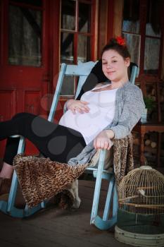 Smiling pregnant woman sitting on a rocking chair on a balcony,  in the countryside