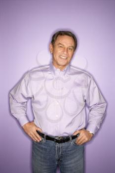 Smiling middle-aged Caucasian man with hands on hips wearing purple clothing on purple background.